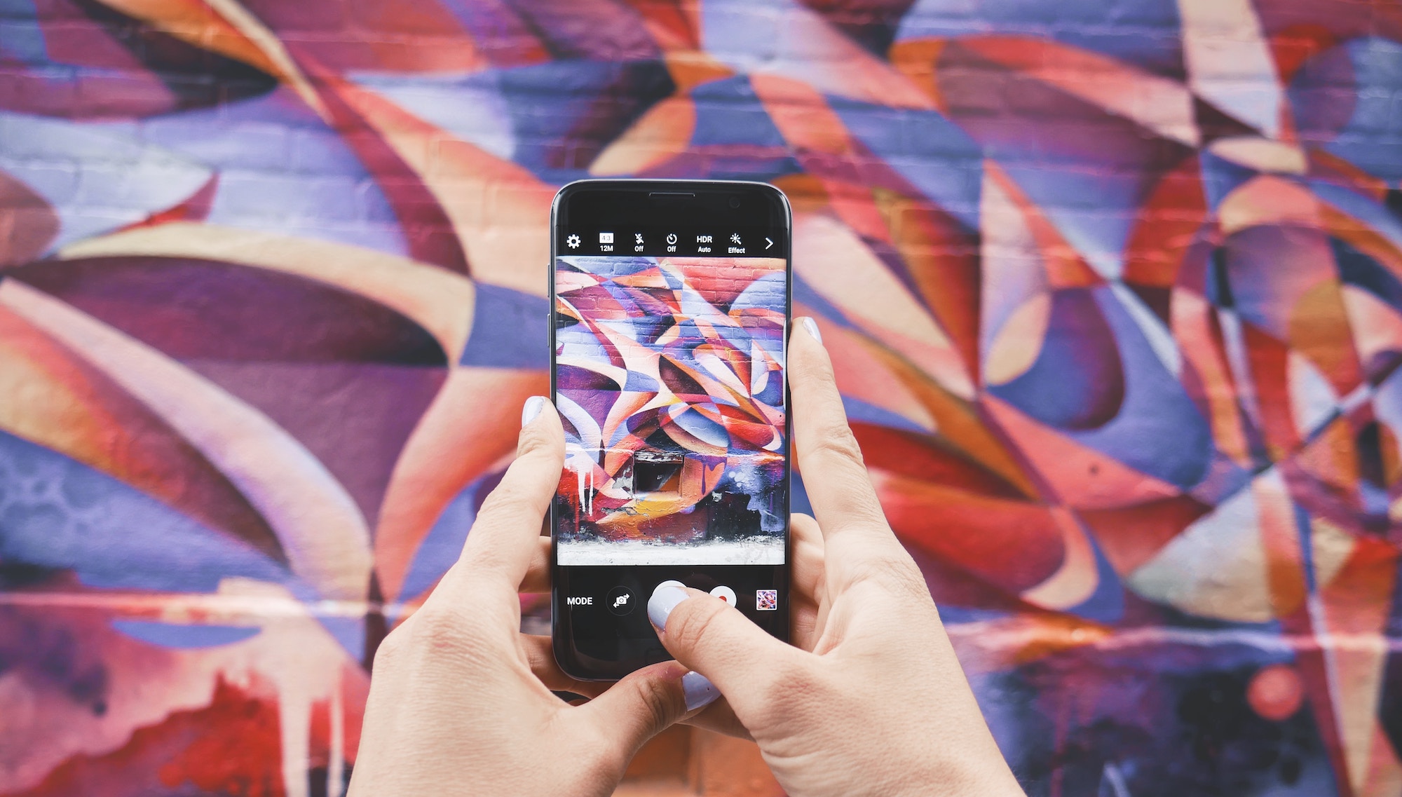 Hands holding a phone up in front of street art - abstract shapes in red, orange and purple