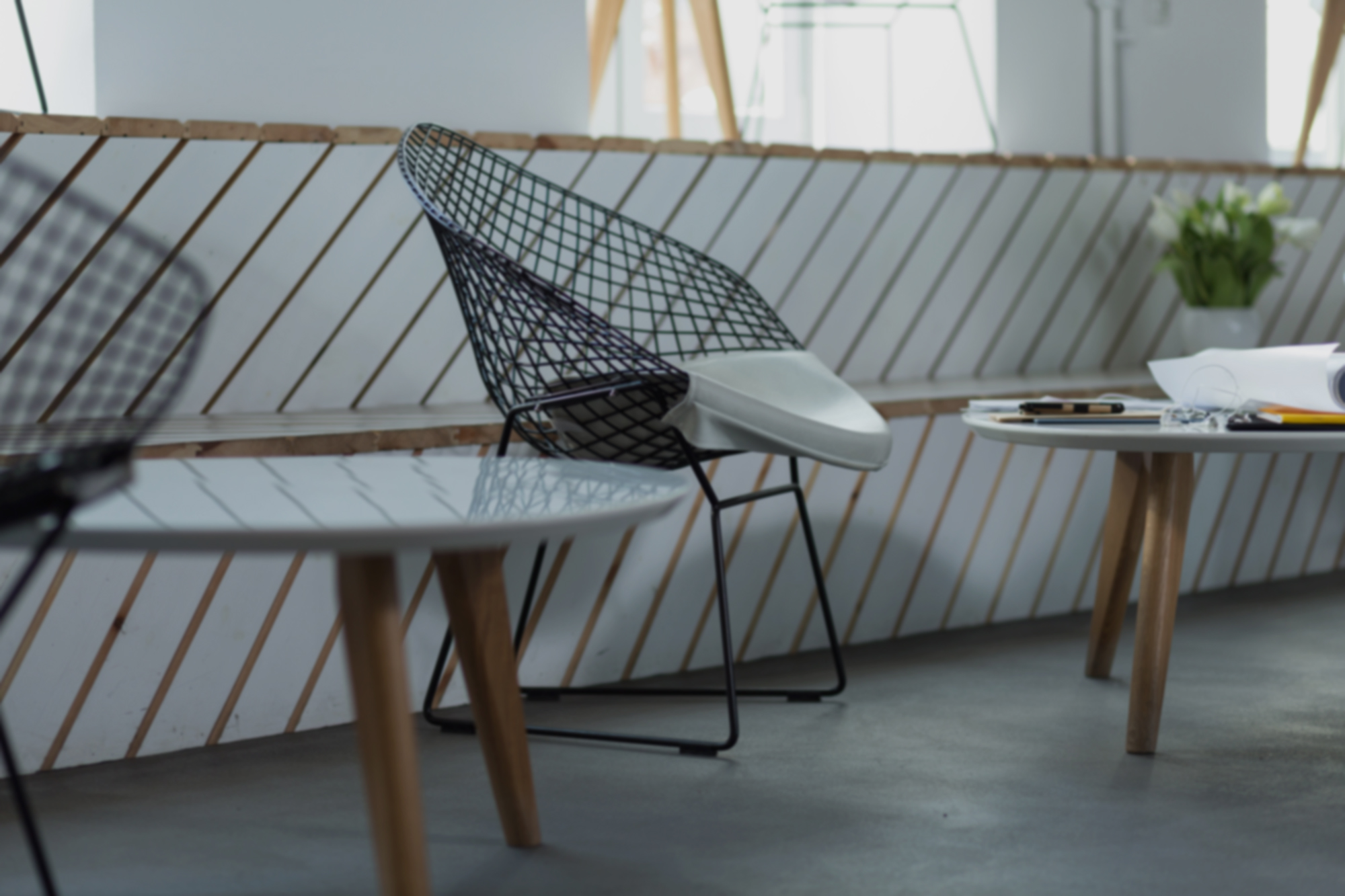 Modern office interior with a wireframe chair and two round tables holding papers, pencils, and plants, suggesting a minimalist creative workspace.