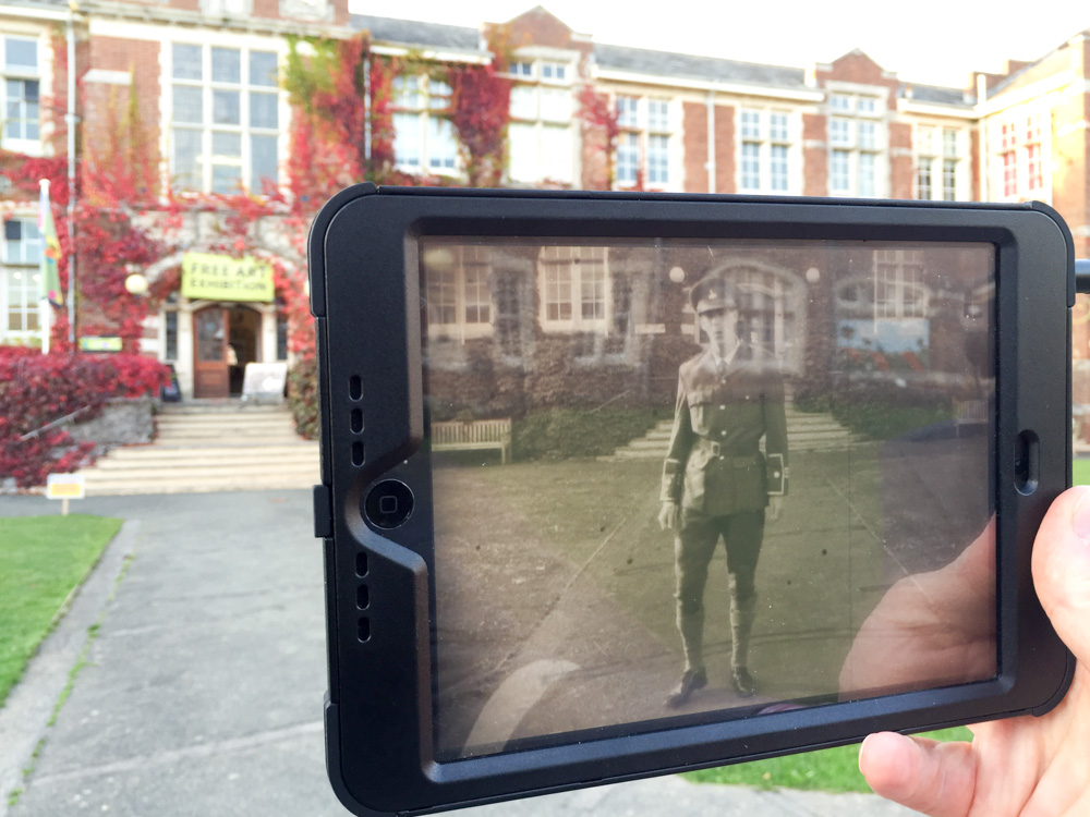 We see an old black and white image of a British military soldier stood outside the hospital on the digital screen of a tablet. The tablet is held up to the hospital, photographed in colour, in present day.