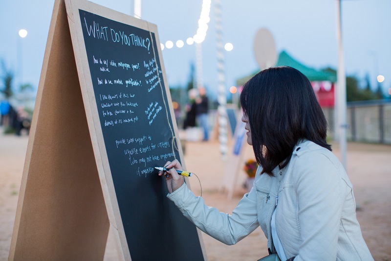 Woman in profile, writing on a chalkboard against a dusk outdoor background with festoon lighting