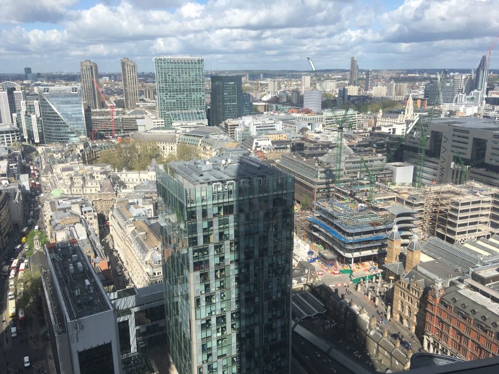 A view over central London in the morning, taken from a tall building, the scene shows glass-windowed high-rise buildings and older architecture below, with construction sites dotted in the background.