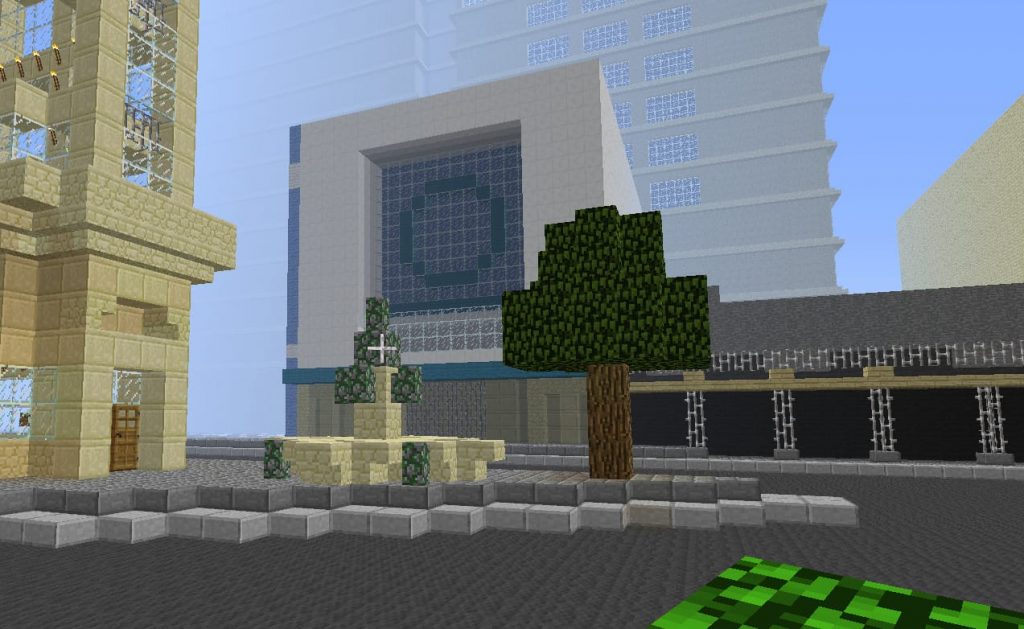 A city street is depicted in MineCraft, with cubic, blocky versions of a road, pavement, fountain, tree and yellow-stoned buildings in the background.