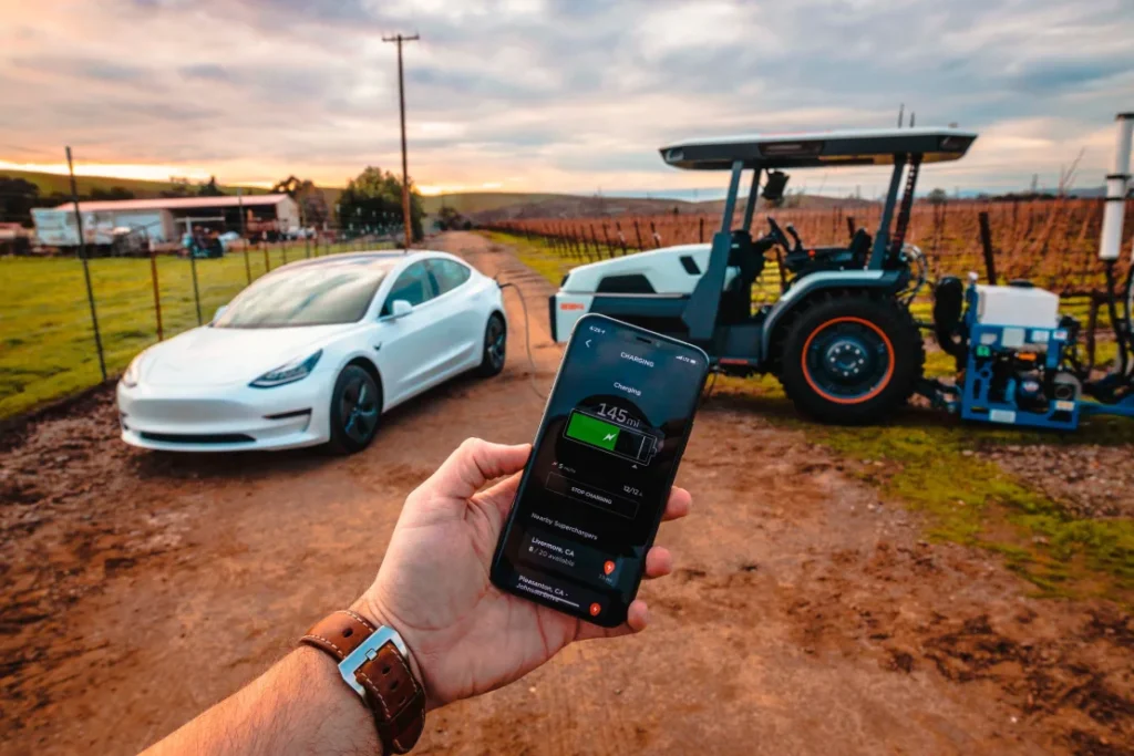 Hand holding a phone which shows tractor charging level. In background there is an electric tractor, an electric car and farm buildings
