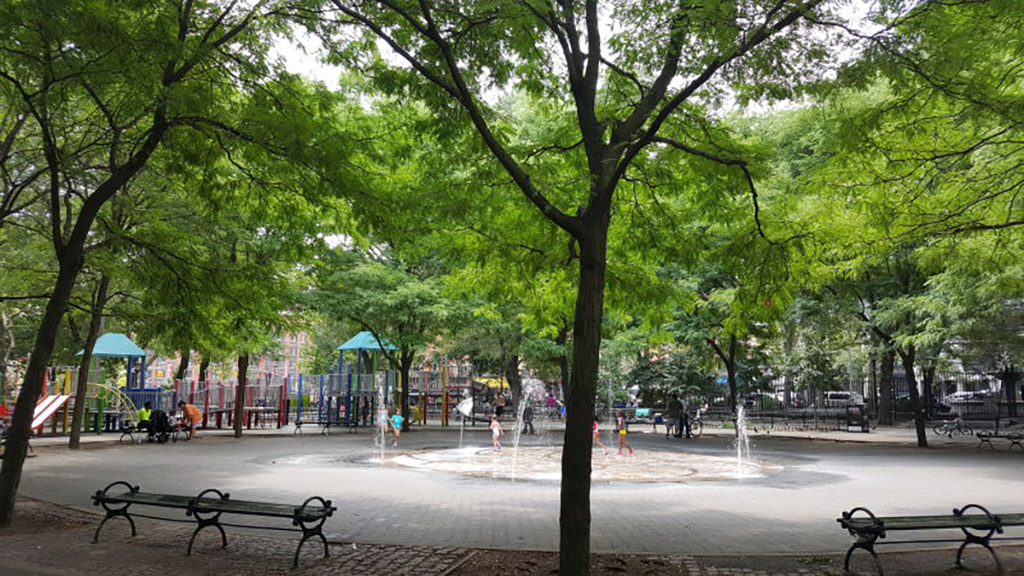 View past trees and benches through to central area with floor level fountains and children playing. Playpark, street and seating in background.