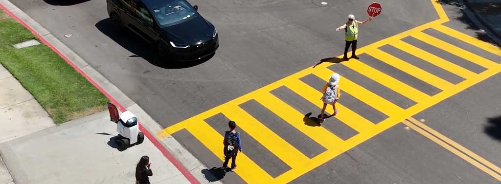 A short, boxy wheeled robot near a kerb edge, next to a pedestrian crossing with children, traffic warden and cars.