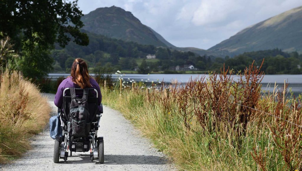 Person in wheelchair on a smooth gravel path, next to a lake surrounded by dramatic hills.