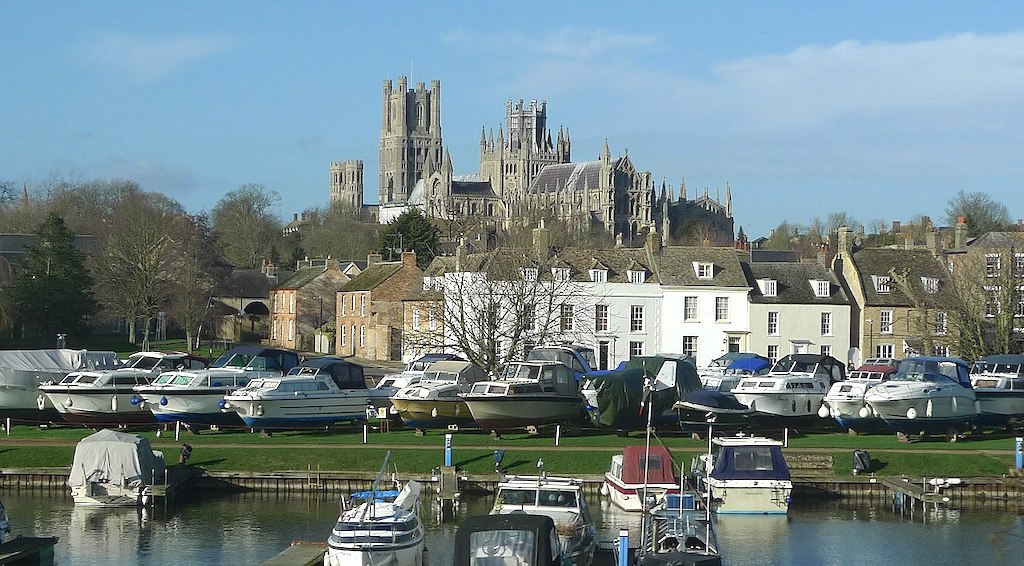 Leisure boats on a river,, with more boats moored on land behind, with a terrace of houses behind them, and Ely Cathedral in the background