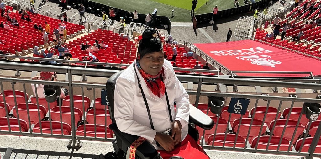 Lady-Marie dressed in white and red, smiling to camera, with the terraces of the Arsenal football club behind, descending to the pitch.