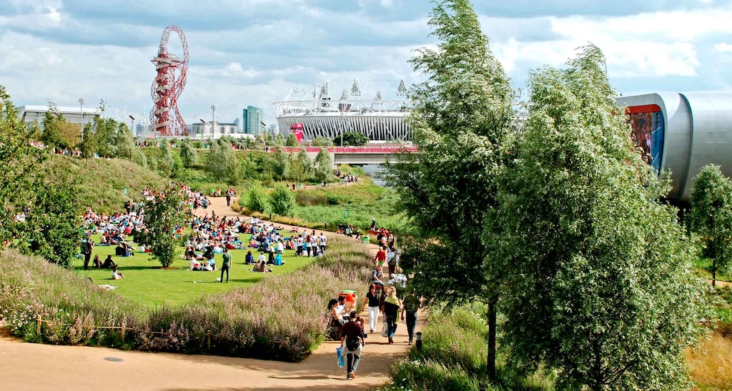 Busy park with paths, landscaping, Olympic buildings on a sunny day.