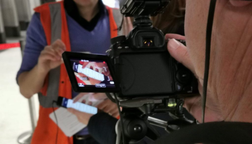 View through a digital camera at a person holding a phone. People in orange hi-vis vests in the background.