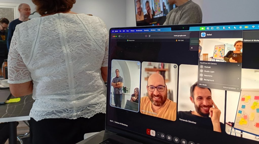 Laptop showing two of the team smiling via a video call, with others in the team standing behind, looking to the video call feed on the wall screen.