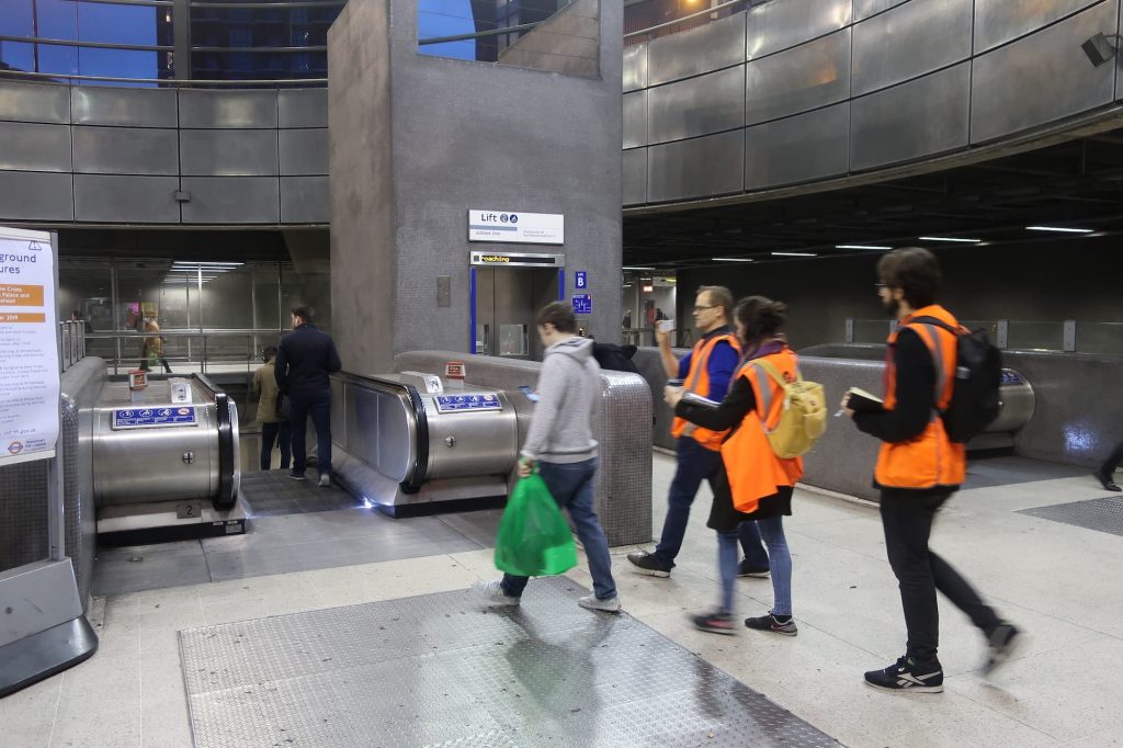 People in a train station. Three people in orange hi-vis jackets are walking behind a person holding a phone.