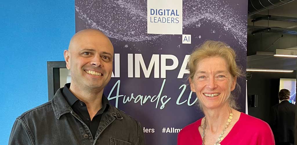 Jo and Adam smiling to camera in front of Digital leaders AI Impact awards banner.