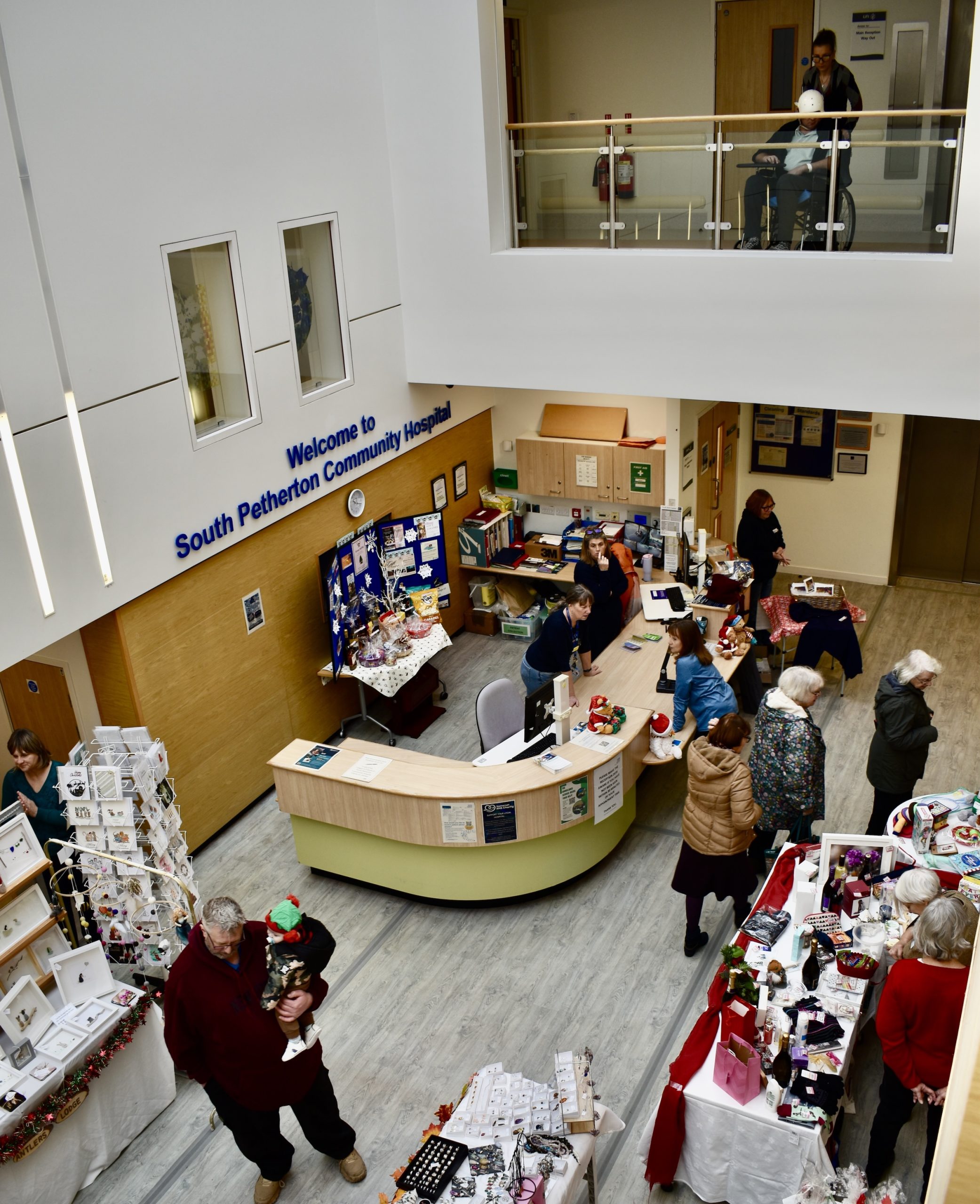 View from community hospital balcony down to reception, with Christmas decorations and craft stalls, staff, patients and visitors.