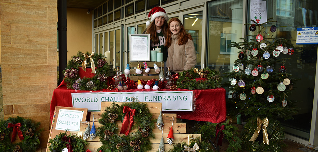 Two people smiling to camera behind a stall selling pine tree-based Christmas decorations to the side of a community hospital entrance.