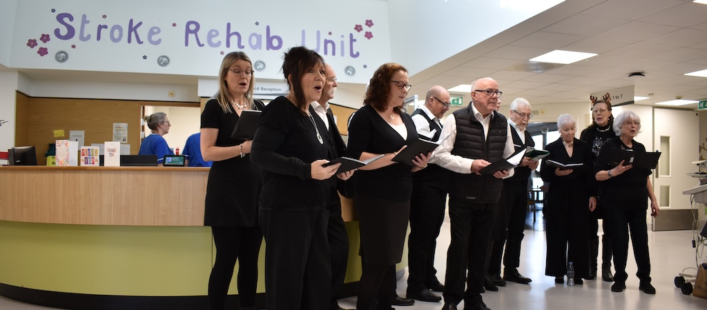 A community choir of 15 people singing, stood in front of the reception desk with nurses working behind.