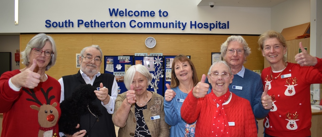 Severn event volunteers smiling and giving 'thumbs up' to camera, in front of the reception sign "Welcome to South Petherton Community Hospital"