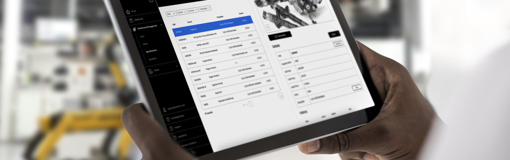 Hands holding a tablet with data in tables, with factory equipment behind.