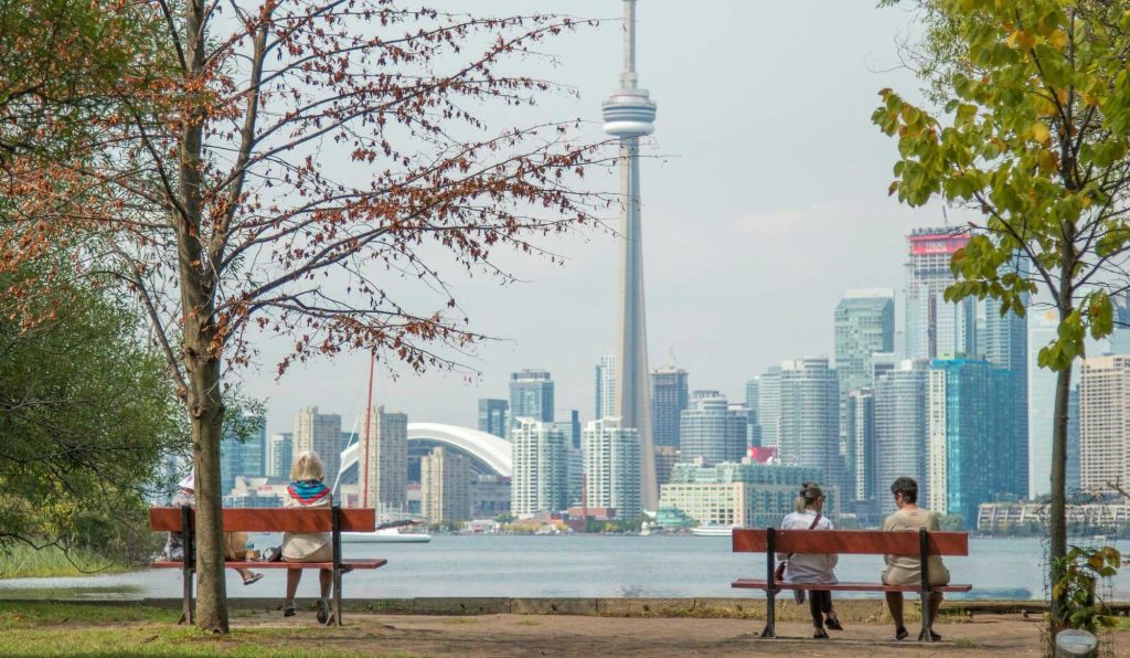 People sat on park benches with trees, looking over the water to the Toronto skyline