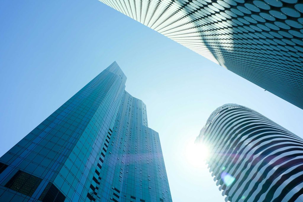 Looking up to skyscrapers and a stark blue sky, with sun behind edge of one tower corner.