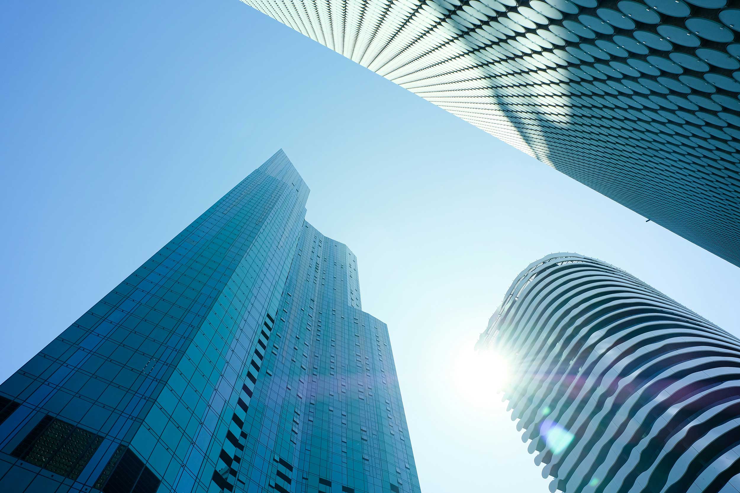 Looking up to skyscrapers and a stark blue sky, with sun behind edge of one tower corner.