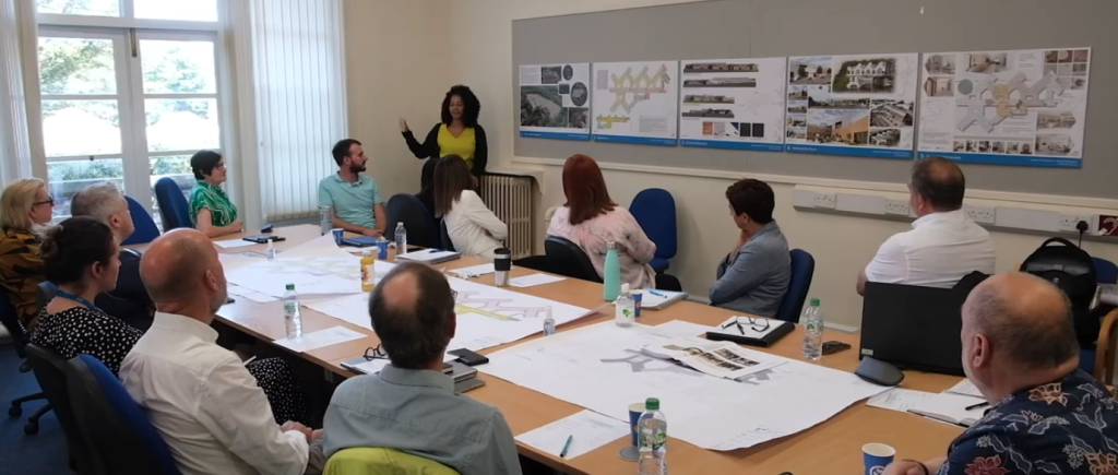 Around 12 people sat around a large table, discussing the plans, maps and drawings on the table and walls.