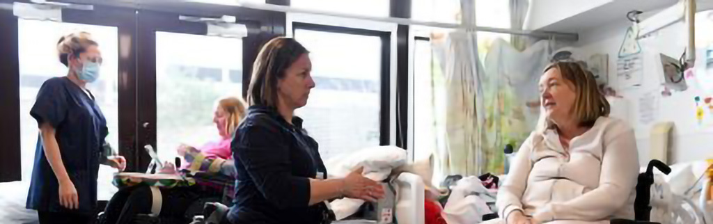Hospital ward with two women with reduced mobility and dexterity in conversation with two nurses.