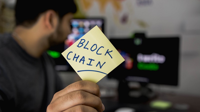 Man holding a sticky-note with the words "Block Chain" written on it, looking at computer screens in the background