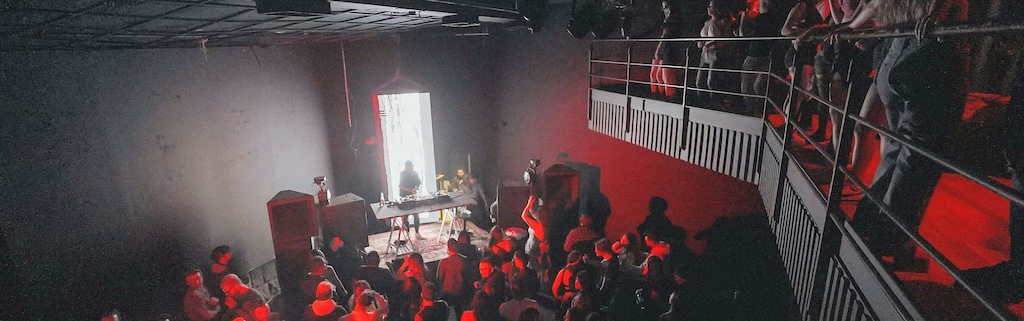 View from a balcony of a DJ on a small stage in a dark club, with bright light from an open door behind. Dancefloor is full, with dancers lit in red light. Balcony is also full with audience.