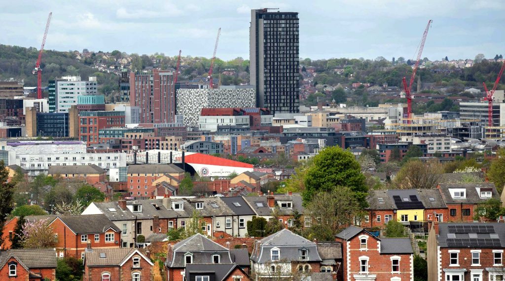 View over roof tops of Sheffield, with cranes, tower blocks, houses and a football stadium.