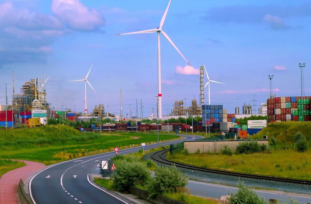 Road through green fields, leading to wind turbines, stacked shipping containers and industrial buildings, under a blue sky.