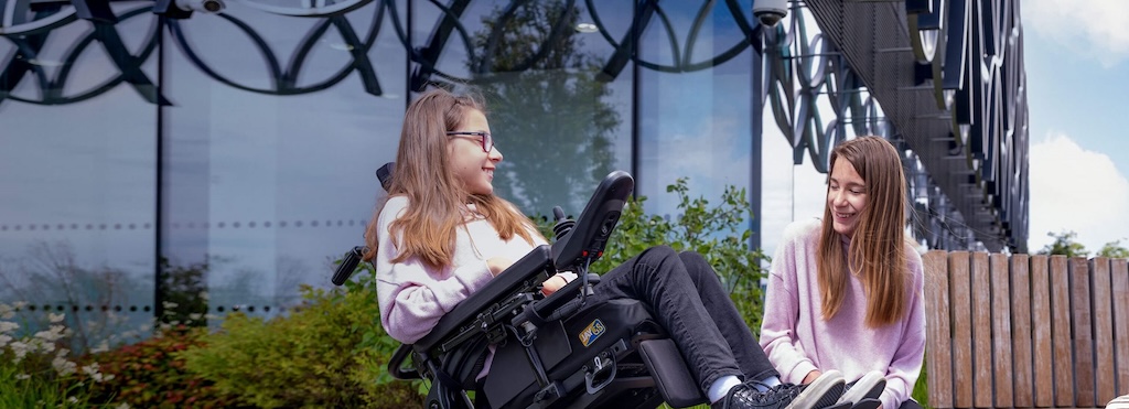 Two teen girls outside smiling and chatting, one in a motorised chair, one on a bench. 