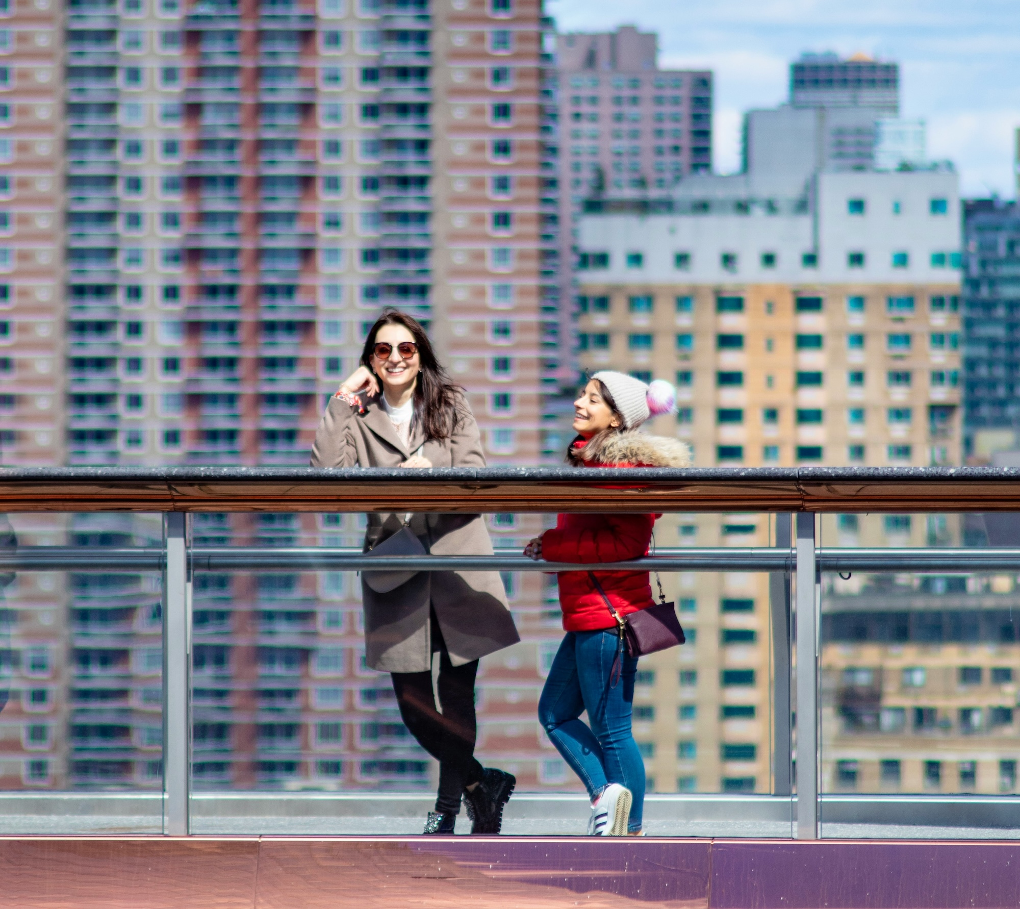 Two women stood on a city roof by a railing, with high rise buildings behind.