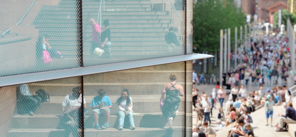 Reflections in a glass-panelled wall of people sat on public steps, and walking down a city park path.