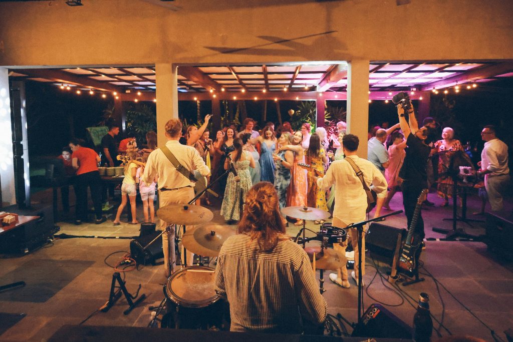 View from behind a drummer in a small band, looking beyond stage to a dancing audience in a small gig venue.