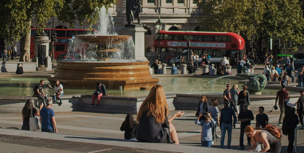 A woman sat on steps in a city square, with walkers, families, a fountain and bus.