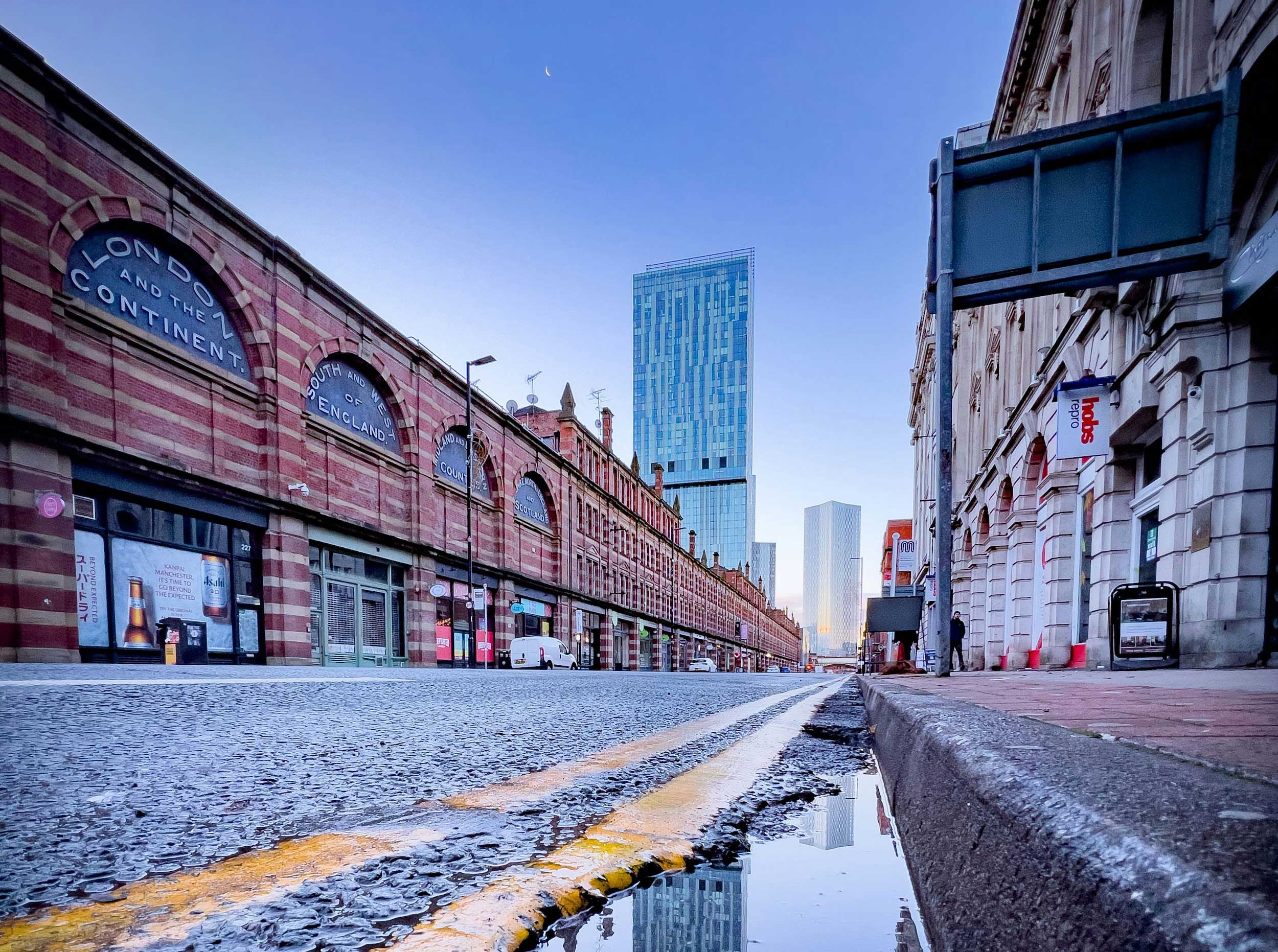 Manchester street from ground level, looking up from puddle to businesses under train arches, to tower blocks in the distance.