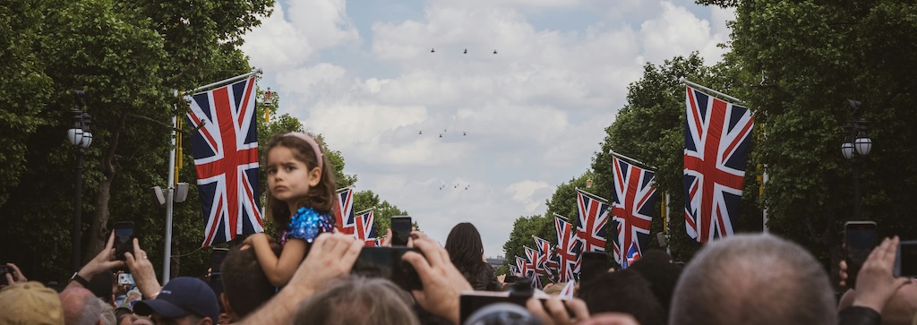 A crowd gathered under Union Jack flags, looking at aerial display above, with a child sat on shoulders turn to look back at the camera.