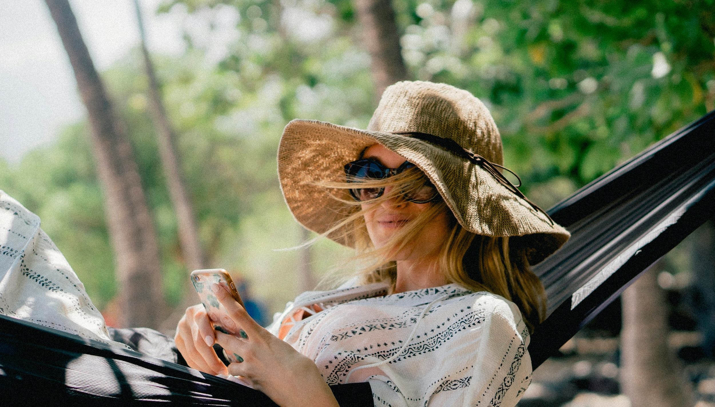 Person in sunglasses and sun hat in a hammock, smiling, and holding a phone.