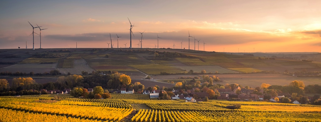 Sunset over crop fields, with a village in the valley beyond, with wind turbines on the hills on the horizon.