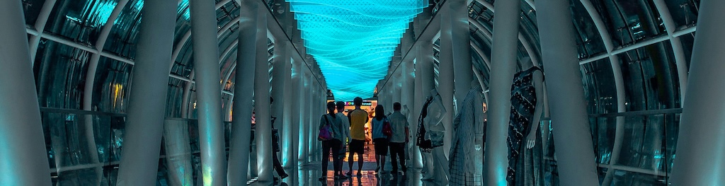 View down a glass skywalk corridor at night, looking towards a group of people walking towards an orange-lit exit.