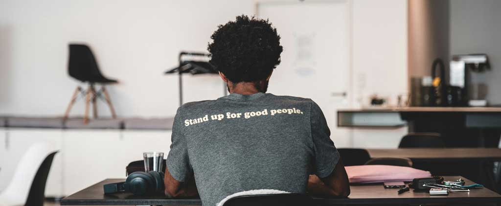 Person at a desk, back to camera. T-short print reads 'Stand up for good people.
