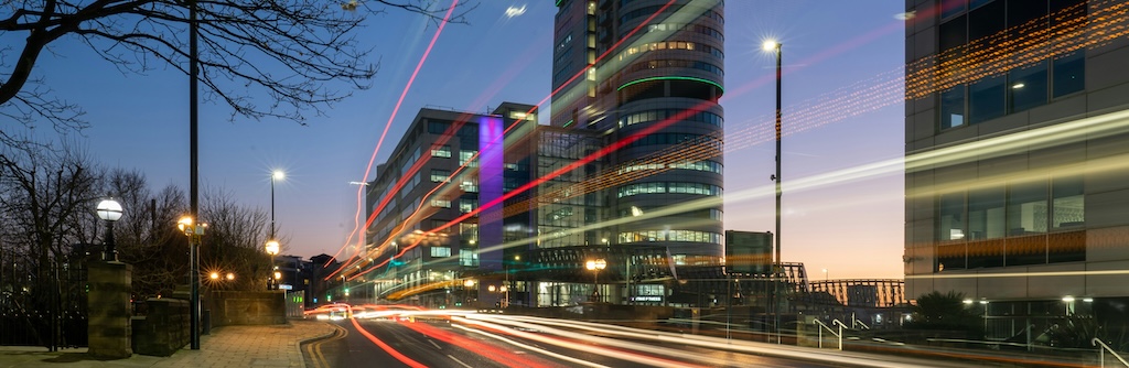 Long exposure night photo of a city, with lights from traffic streaking past tower blocks and a dusky sky. 