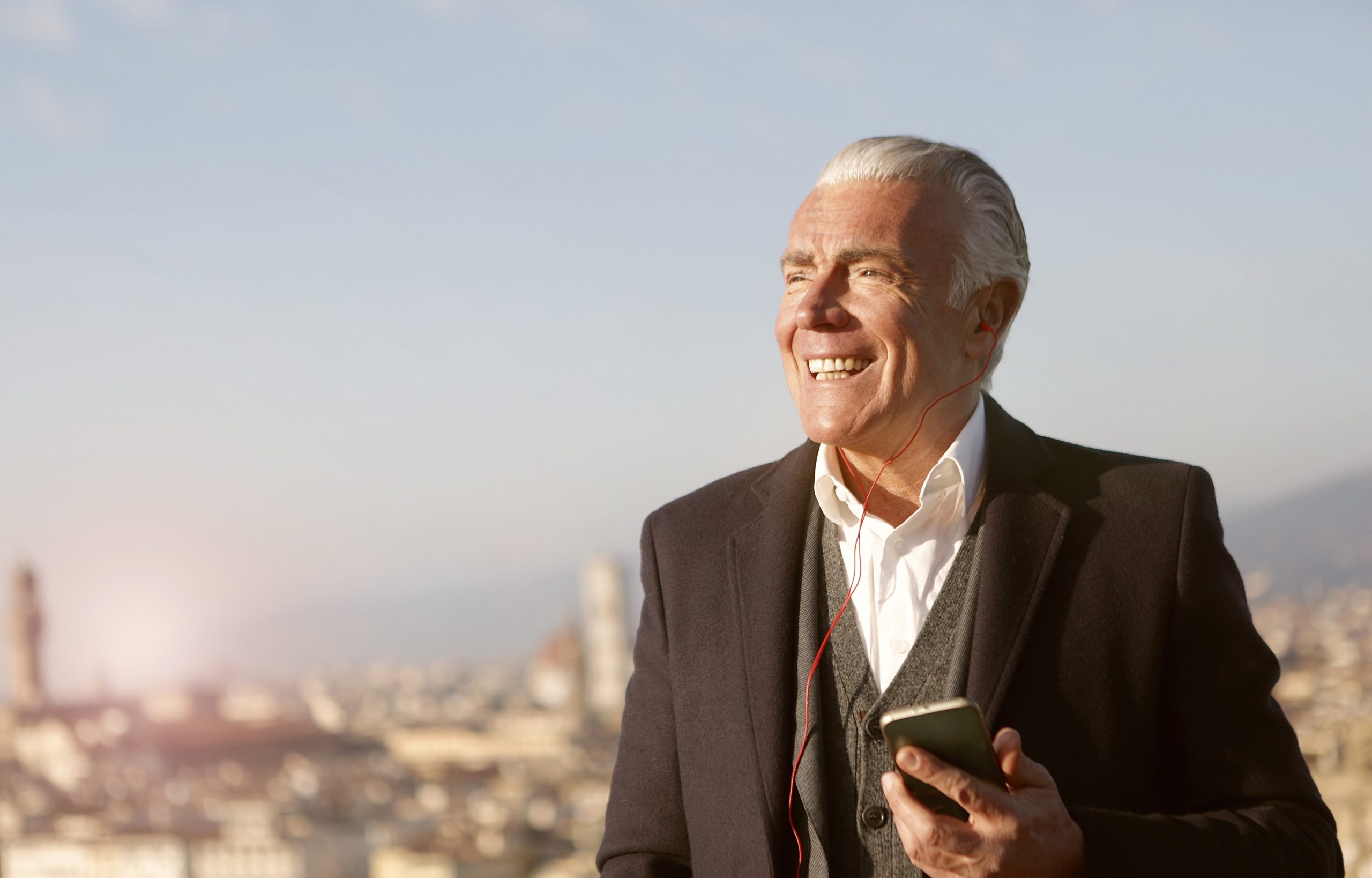 A white man, with grey white hair, in his 70's, smiles as he holds a phone and has headphones in as he listens to a Hidden Cities audio guide with the city skyline behind him