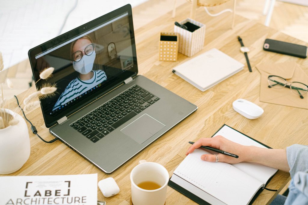 a person is seen having a virtual meeting with the person on the screen wearing a face mask and glasses. The meeting is on a laptop that is on a wooden table and there are notebooks and a mug of coffee on the table.