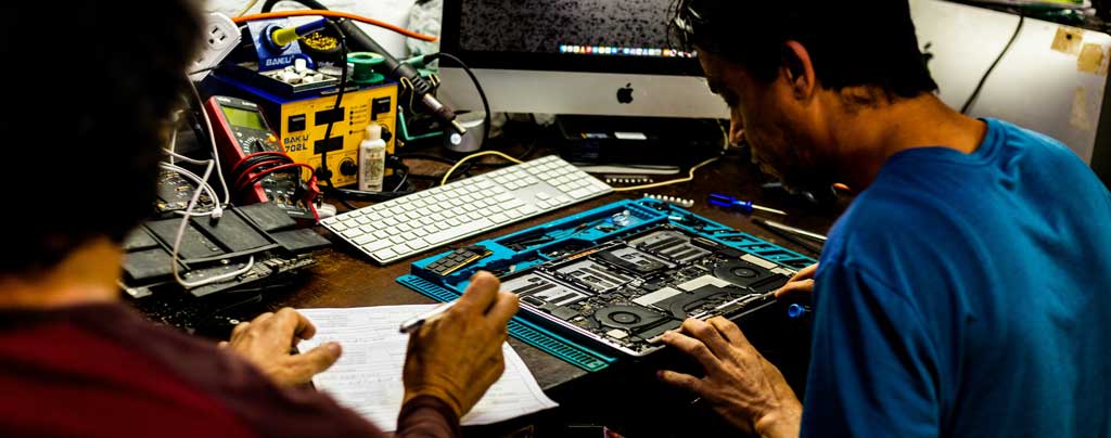 Two people at a desk, one repairing a laptop, the other with a paper form and pen.