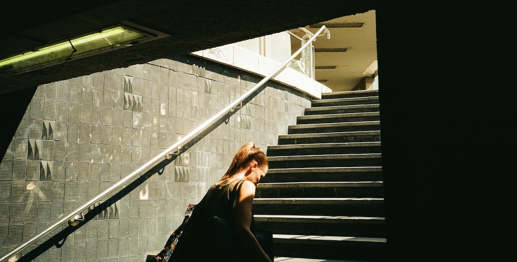 A woman walking up steps from a dark underpass to a sun lit street.