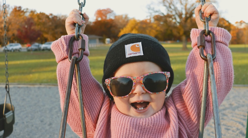 Toddler wearing sunglasses, laughing in a playground swing in a park.