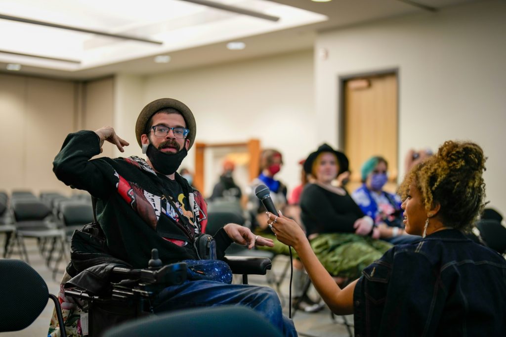 Person with physical impairments talking to a person holding a microphone, in a busy room at an innovation and inclusive design research event.