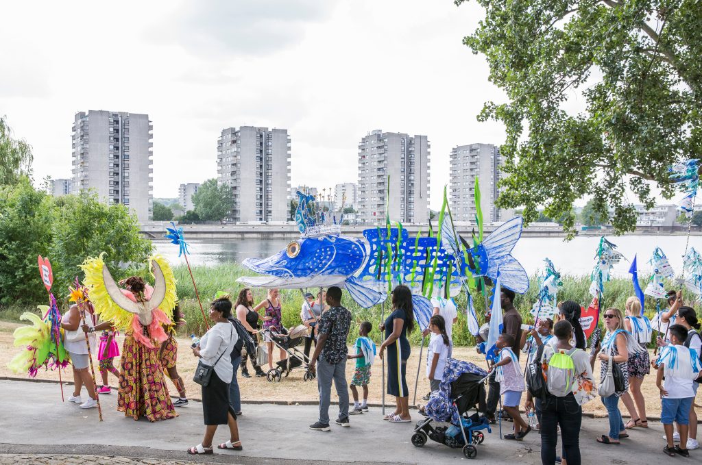 Community procession carrying a large blue fish model and decorations along a road by a lake, with tower blocks across the water in the distance.